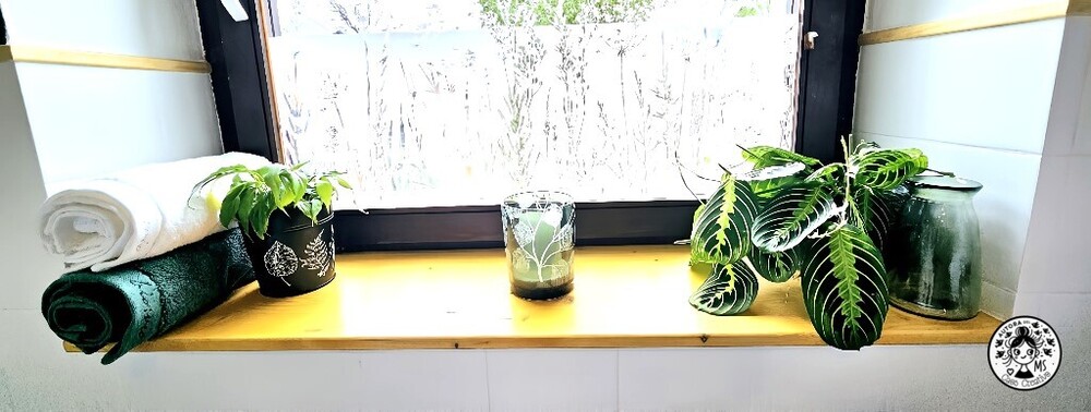 Decoración de repisa en aseo pequeño con toallas enrolladas, plantas naturales y tarro de cristal verde.